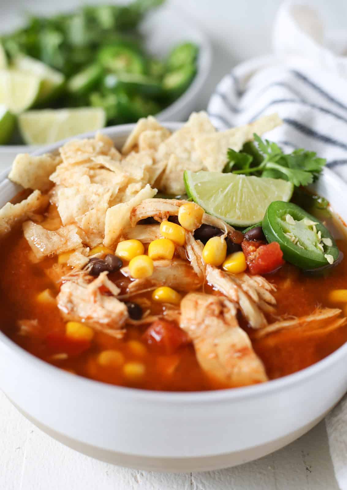Chicken tortilla soup in a white bowl topped with crushed tortilla chips, cilantro, fresh jalapenos, and a lime wedge.