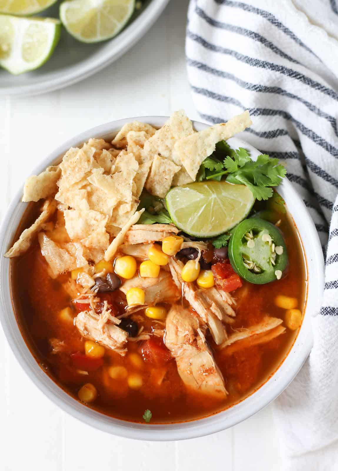 overhead shot of a bowl of chicken tortilla soup topped with crushed tortilla chips, cilantro, jalapeno slices, and a lime wedge